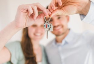 Un couple en arrière-plan tenant un jeu de clés avec un porte-clé en forme de maison au premier plan